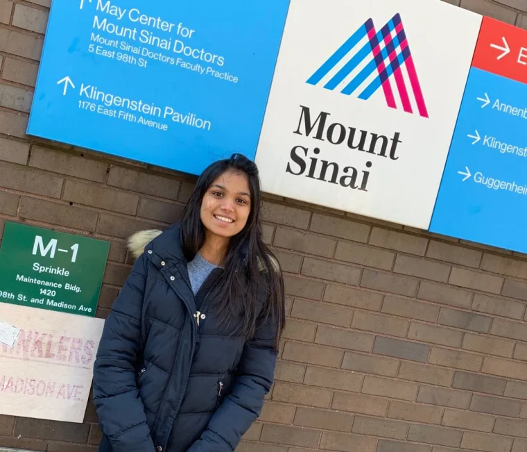 Person in a black coat stands smiling in front of a brick wall with a Mount Sinai sign.