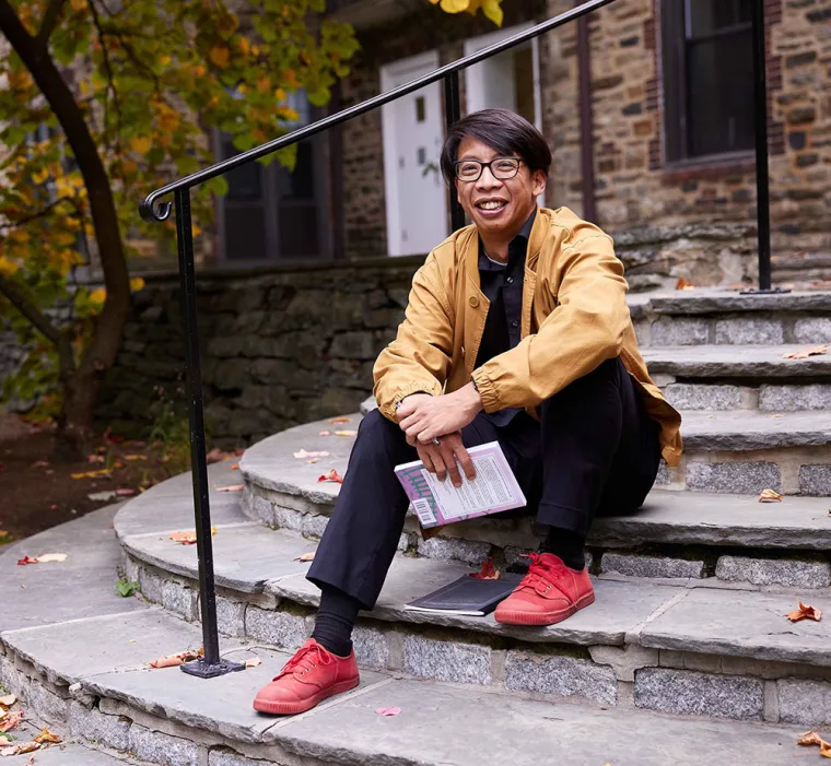 Person sitting on stone steps, holding a book, smiling. Wearing a tan jacket, black pants, and red shoes, with autumn leaves around.