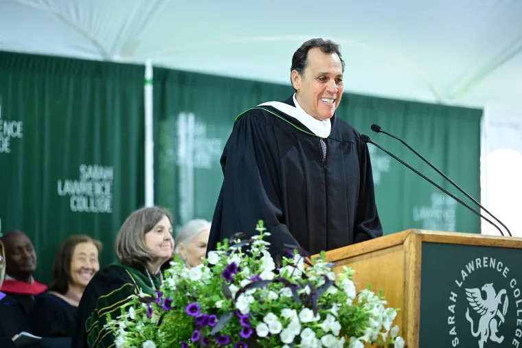 Sam Edelman speaking at a podium with "Sarah Lawrence College" banners in the background. Flowers are arranged in front.