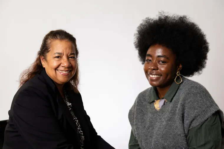 Two individuals are sitting and smiling at the camera. One wears a suit jacket while the other wears a grey sweater with a collared shirt.