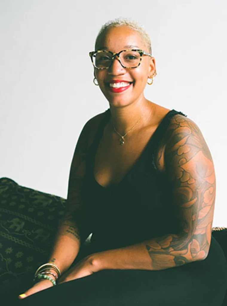 Person with short hair, glasses, and tattooed arm smiles while sitting on a patterned surface, wearing a black sleeveless top.