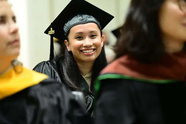 Graduate in cap and gown smiling, surrounded by others in similar attire at a ceremony.