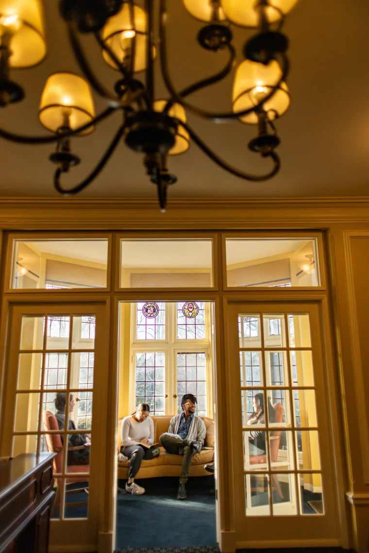 Four people sit and converse in a warmly lit room with large windows and a chandelier.