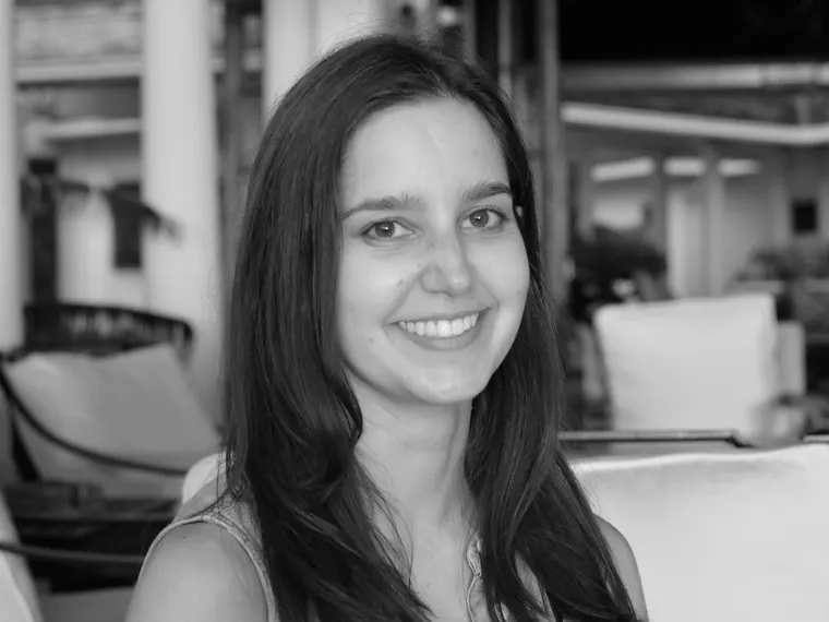 Person with long hair smiling, seated indoors on a sofa, with soft-focus background elements like chairs and columns. Image in black and white.