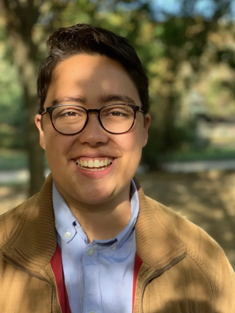 Person with glasses smiling outdoors, wearing a brown jacket and blue shirt. Sunlight creates dappled shadows on their face.