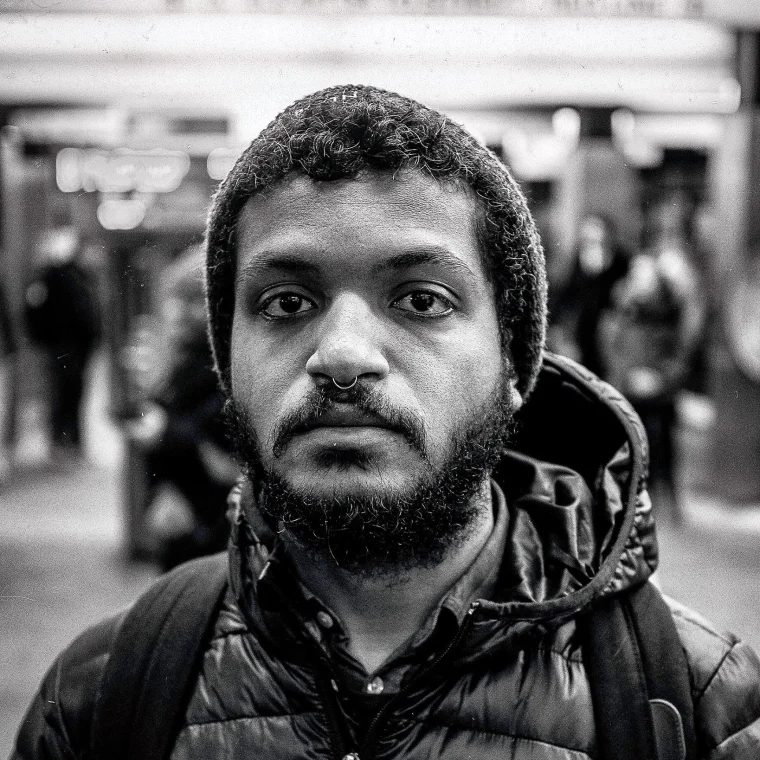 Person with a beard and nose ring wearing a beanie and puffy jacket, standing in a busy indoor setting. Black and white image.