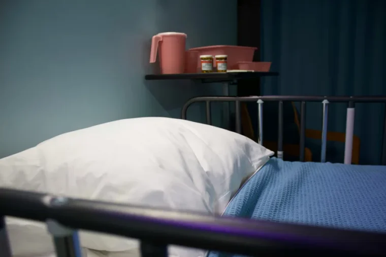 Hospital bed with a white pillow and blue blanket. A nearby shelf holds a pink jug, tray, and medicine bottles in a dimly lit room.