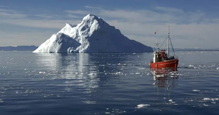 Red fishing boat in icy waters with a large iceberg in the background under a clear sky.