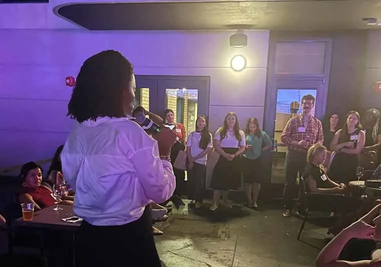 Person speaking into a microphone while a group of people listens attentively in a dimly lit room.