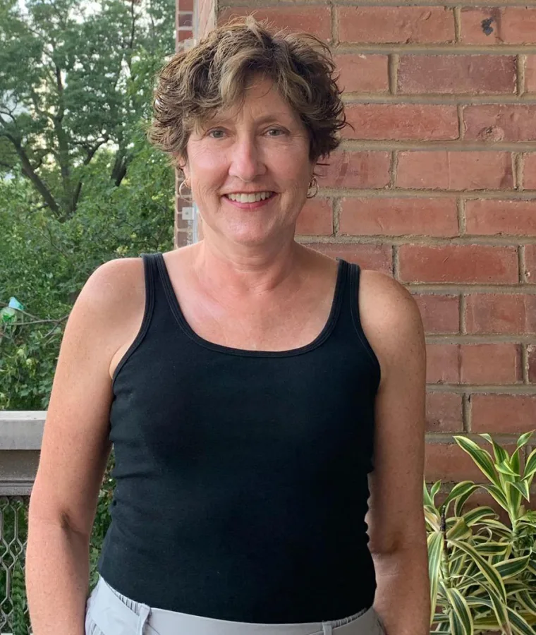 A person with short curly hair smiles in front of a brick wall and trees, wearing a black tank top and light-colored pants.