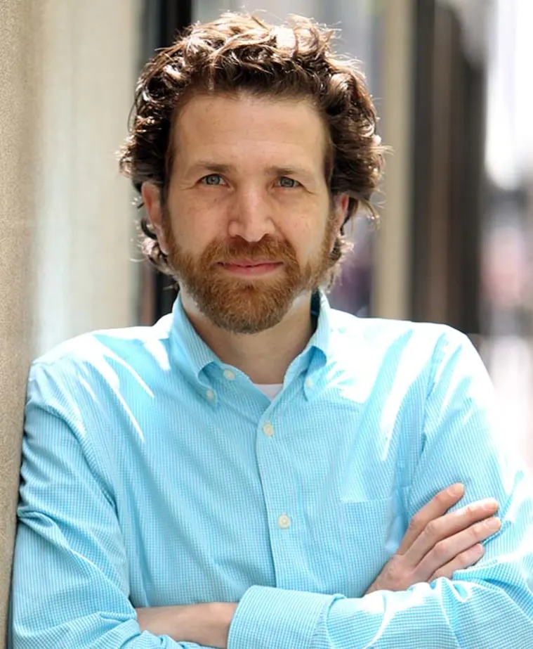 Person with curly hair and beard, wearing a light blue button-up shirt, leaning against a wall with arms crossed, looking at the camera.