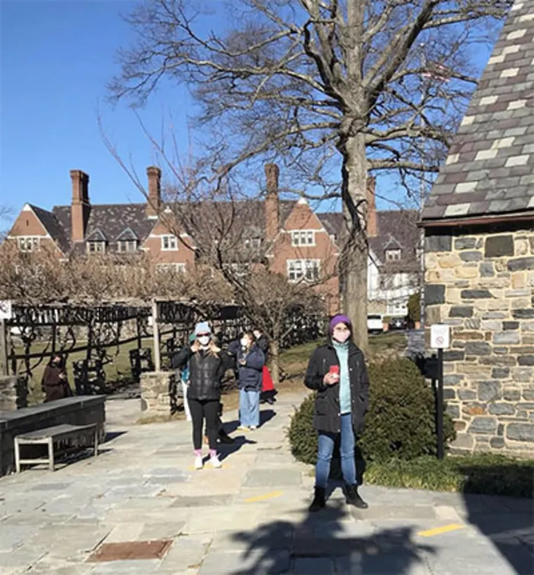 People standing outside on a sunny day, wearing masks, near stone buildings and trees. There's a clear blue sky in the background.