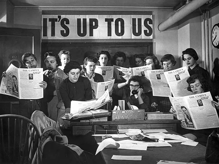 Group of people reading newspapers titled "The Campus" around a cluttered table, with a sign overhead saying "IT'S UP TO US."