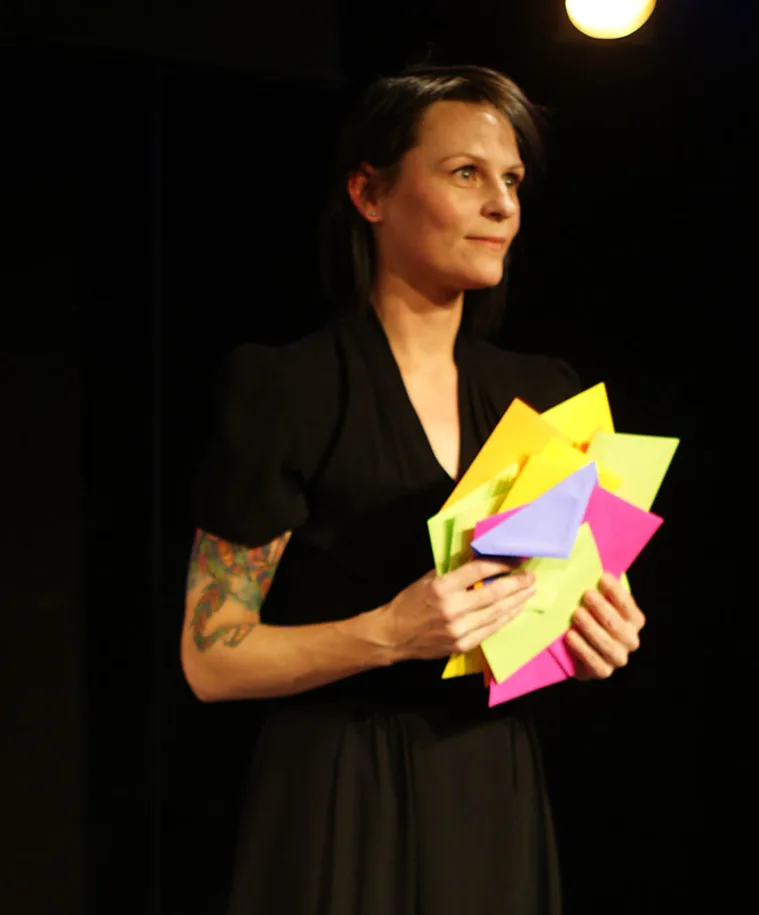 Person in black holding colorful envelopes, standing under a bright light, with a tattoo visible on one arm.