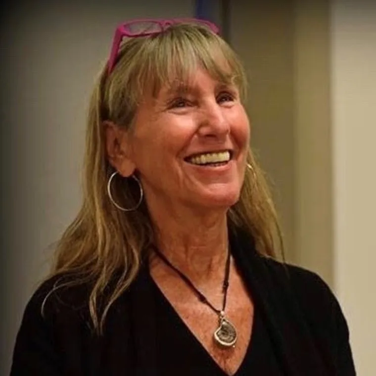 Smiling person with long hair and hoop earrings, wearing pink glasses on head and a pendant necklace, in a warmly lit room.