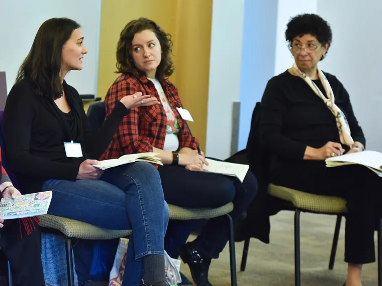 People in a group discussion, sitting on chairs with notebooks. One person is speaking, while others listen attentively.