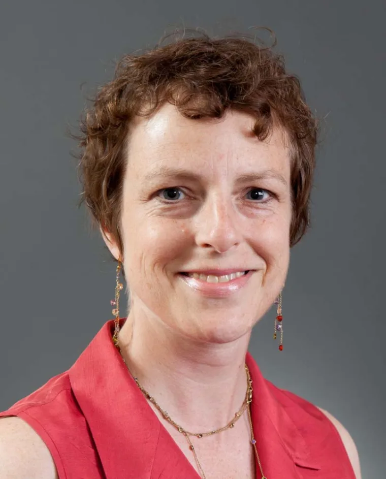 Person smiling with short curly hair, wearing a sleeveless red top, gold necklace, and dangling earrings against a gray background.