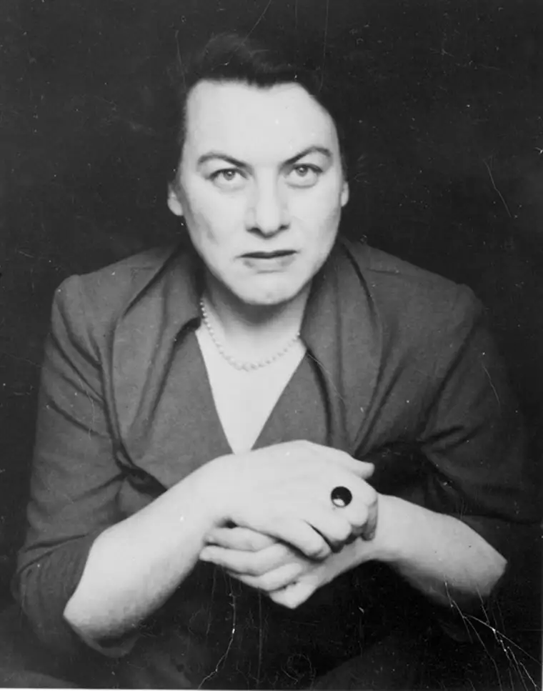 Person in dark clothing with a pearl necklace, looking into the camera with hands clasped.