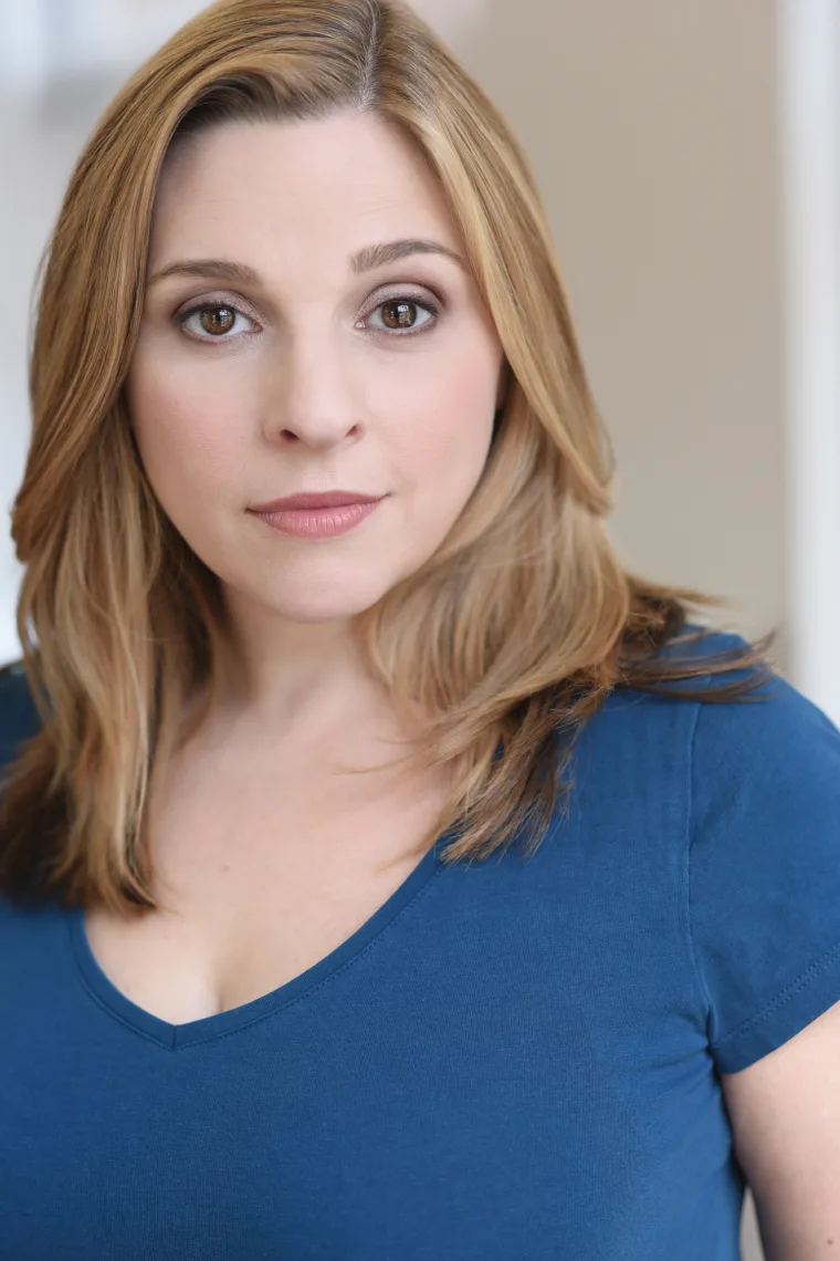 Person with medium-length blond hair wearing a blue top, looking directly at the camera with a neutral expression.
