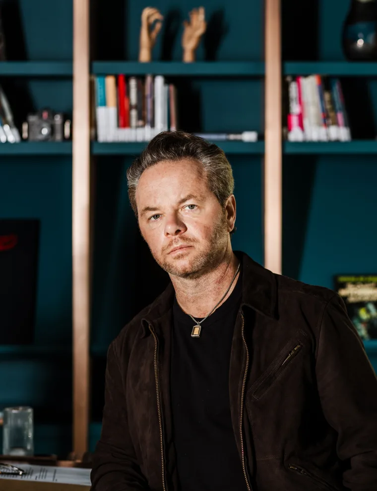 Person with short hair in a dark jacket, sitting in front of a bookshelf with various books and decorative items.