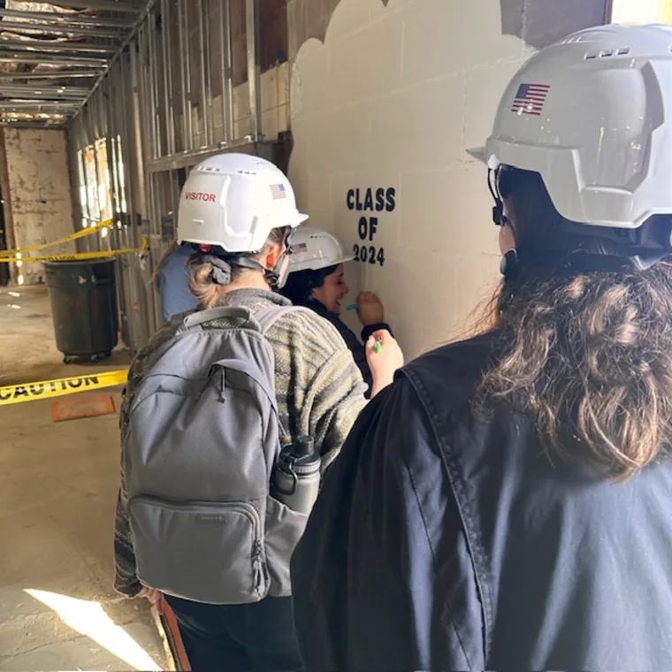 Individuals in hard hats signing a wall that reads "Class of 2024" in a construction area with caution tape. One has a backpack.
