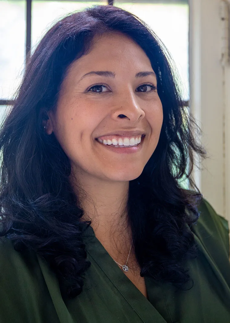 Person with long dark hair smiling, wearing a green shirt, in front of a window.