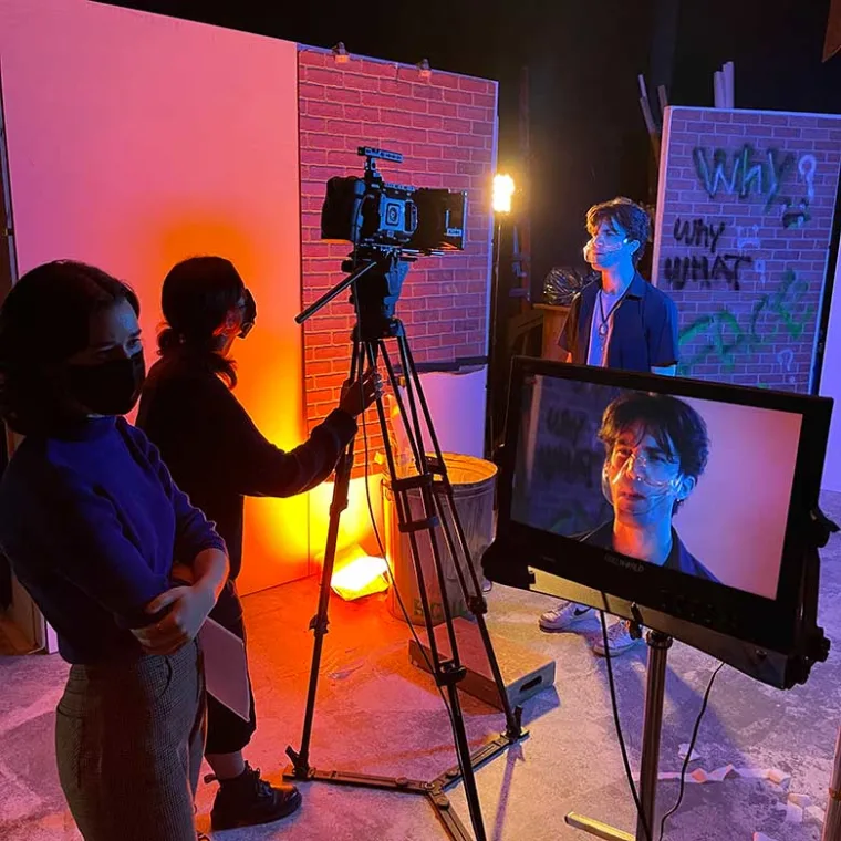 Film set with a camera crew. A person stands in front of a camera, visible on a monitor. Graffiti on the walls and orange lighting.