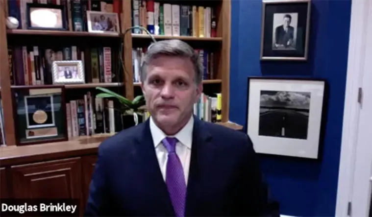 Person in a suit and tie in an office with bookshelves, two framed photos, and a plant.