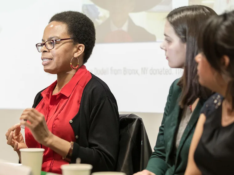 A person speaks at a panel discussion while three others listen, with a blurred presentation in the background.