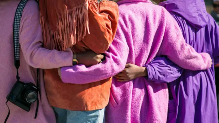 Four people, dressed in pink and purple coats, stand side by side, embracing each other with a camera hanging from one shoulder.