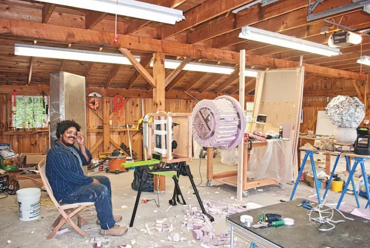 Person sitting in a wooden workshop surrounded by tools and sculpting materials, with large round object on a stand and debris on the floor.