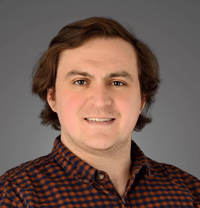 Person smiling with medium-length dark hair, wearing a checkered shirt, against a gray background.