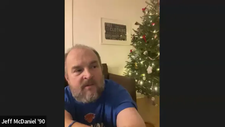 Person with a beard in a blue shirt sits near a decorated Christmas tree. A name tag "Jeff McDaniel '90" is visible on the left.