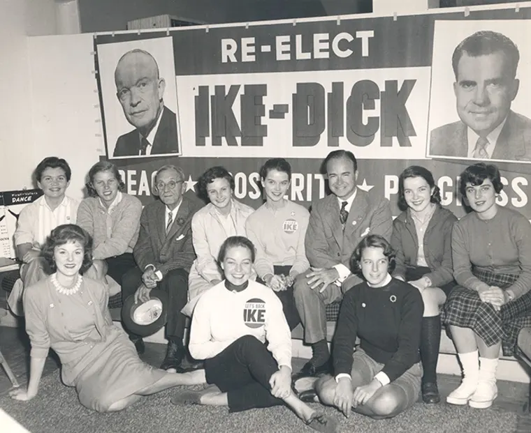 Group of people sitting and kneeling in front of a large "Re-elect Ike-Dick" campaign sign, with two portraits above.