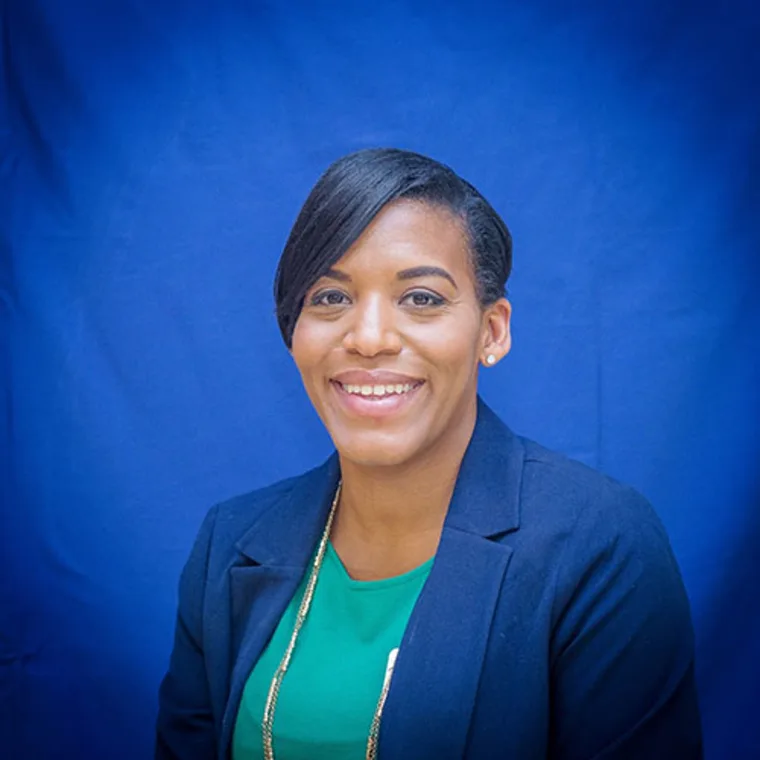 Smiling person wearing a green shirt and dark blazer, in front of a blue background.
