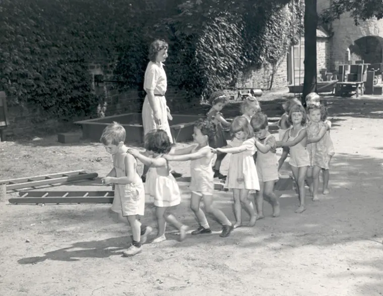 Children are walking in line with hands on each other's shoulders in an outdoor setting with a supervising adult nearby.