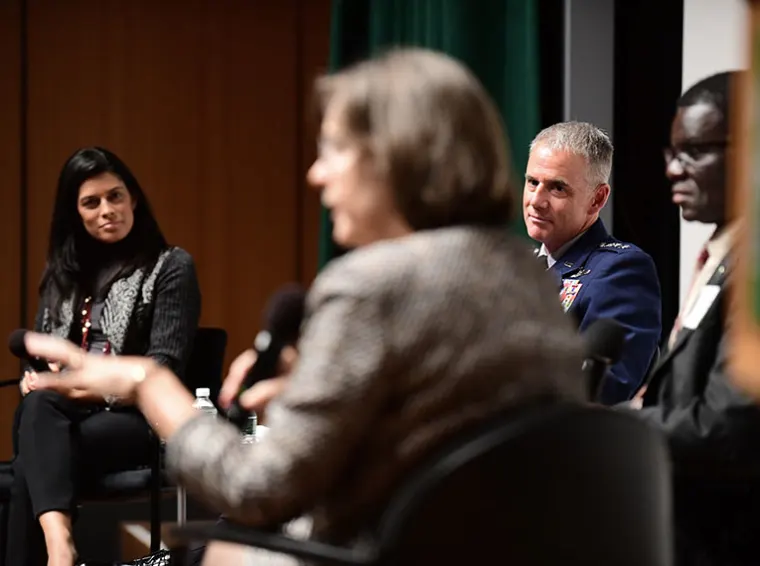 Four people sit in a panel discussion. One person speaks into a microphone while others listen. The background is dark with a green drape.