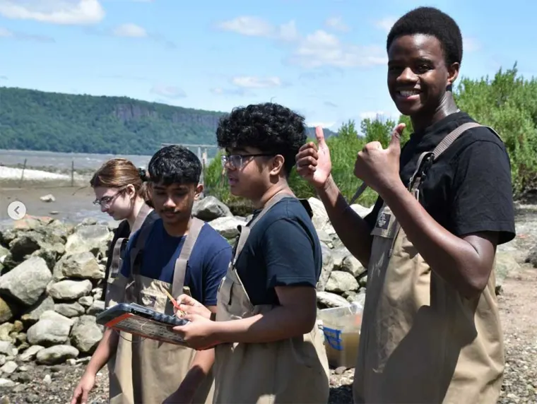 Four people in waders stand by a rocky shoreline. One holds a clipboard and another gives a thumbs-up. Trees and hills are in the background.