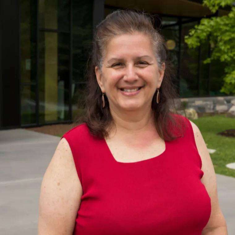 Person in a red top smiles outside near a modern building with large windows and leafy green trees.