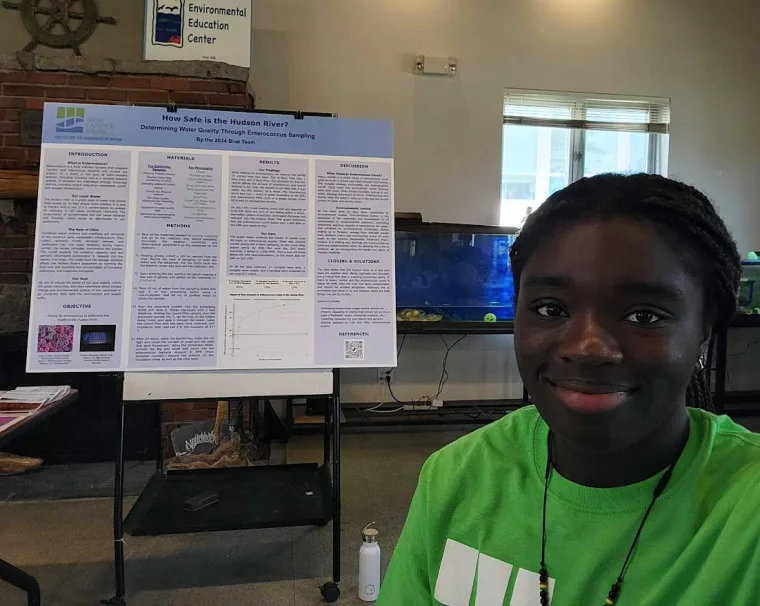 Person in a green shirt smiling in front of a science project poster titled "How Safe is the Hudson River?" in a classroom setting.