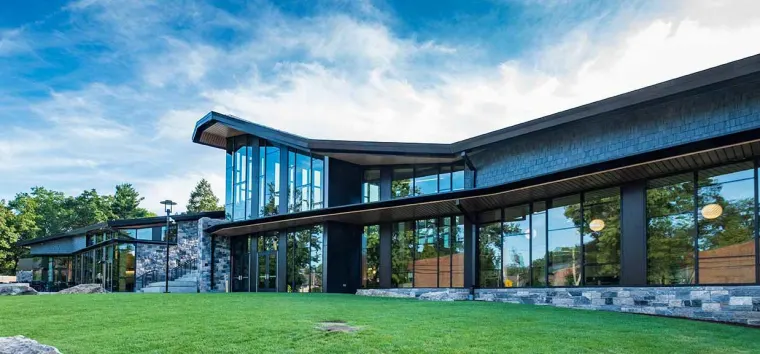 Modern building with large glass windows, angular roof, and stone walls surrounded by green grass and trees under a blue sky.