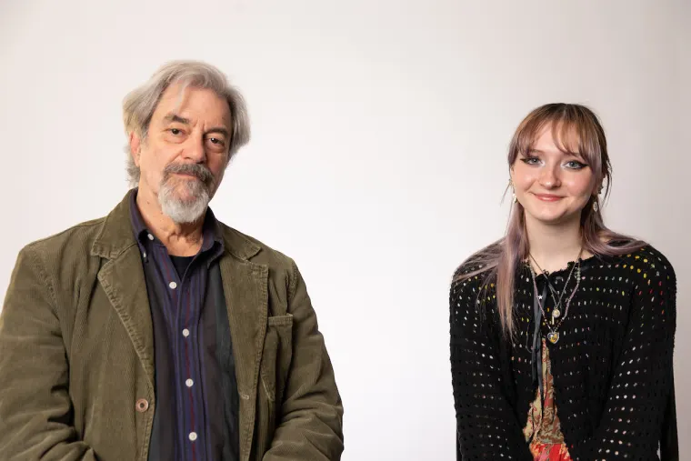 Two people are smiling against a plain background. One is wearing a jacket and shirt, and the other is in a patterned top with long hair.