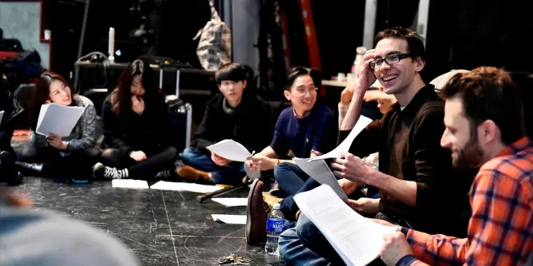 A group of people sitting in a circle on a stage, holding scripts and smiling during a rehearsal or discussion.