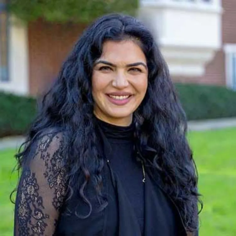 Person with long, wavy hair smiling outdoors with a blurry background of grass and a building.