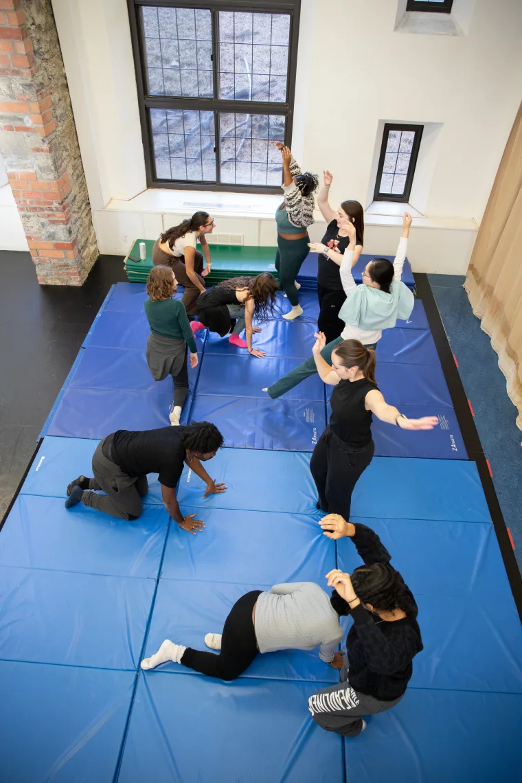 A group of people engaged in a movement exercise on blue mats in a well-lit room with windows and brick walls.