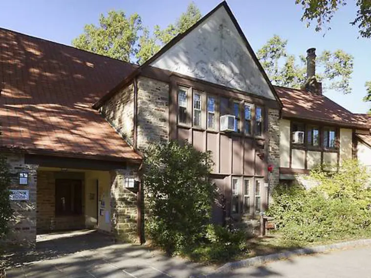 Stone building with brown roof and wooden accents. Arched entrance on the left. Surrounded by greenery and trees under a clear blue sky.