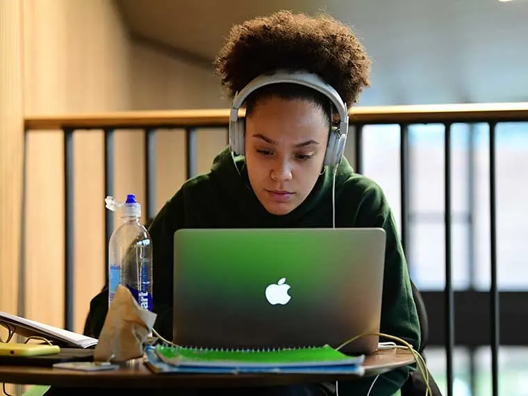 Person wearing headphones intently uses a laptop at a table with notebooks and a water bottle.