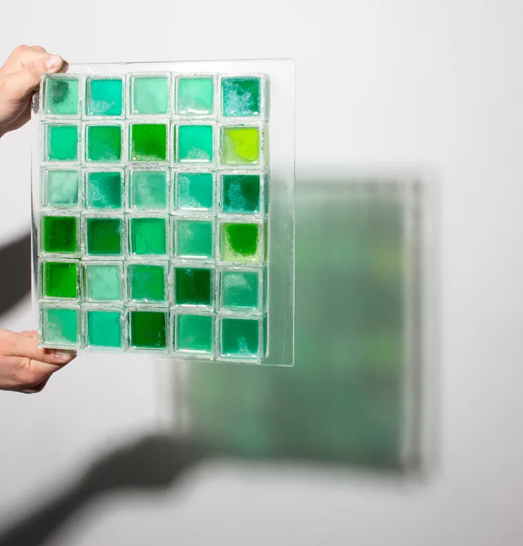 Hands holding a panel of glass tiles in various green shades against a white background.