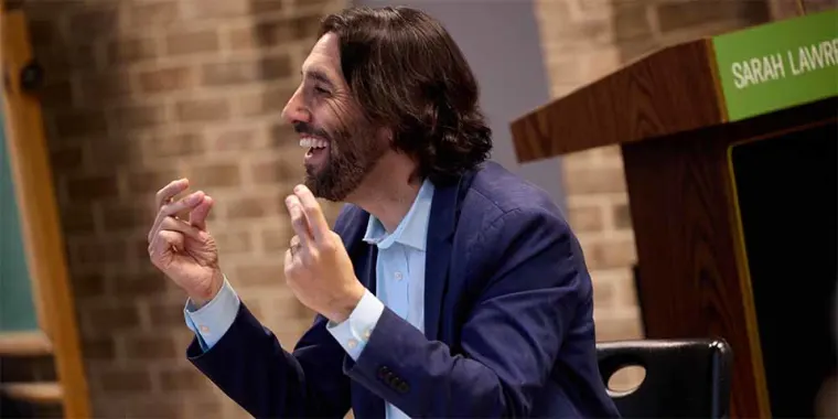 A person with shoulder-length hair and a beard speaks animatedly, sitting near a podium with "Sarah Lawrence" visible in the background.