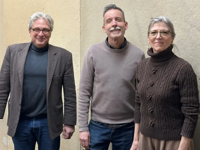 Three people stand in front of a beige wall, dressed in sweaters and jackets, smiling at the camera.